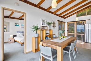 A modern interior with a wooden dining table, white chairs, a kitchen area, and a bedroom with a bed and bedside table.