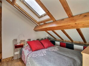 A bedroom with a sloped ceiling, wooden beams, a skylight, a bed with red and grey bedding, and a bedside table with a lamp. - Spacious duplex for 8 near the slopes (La Plagne 1800)