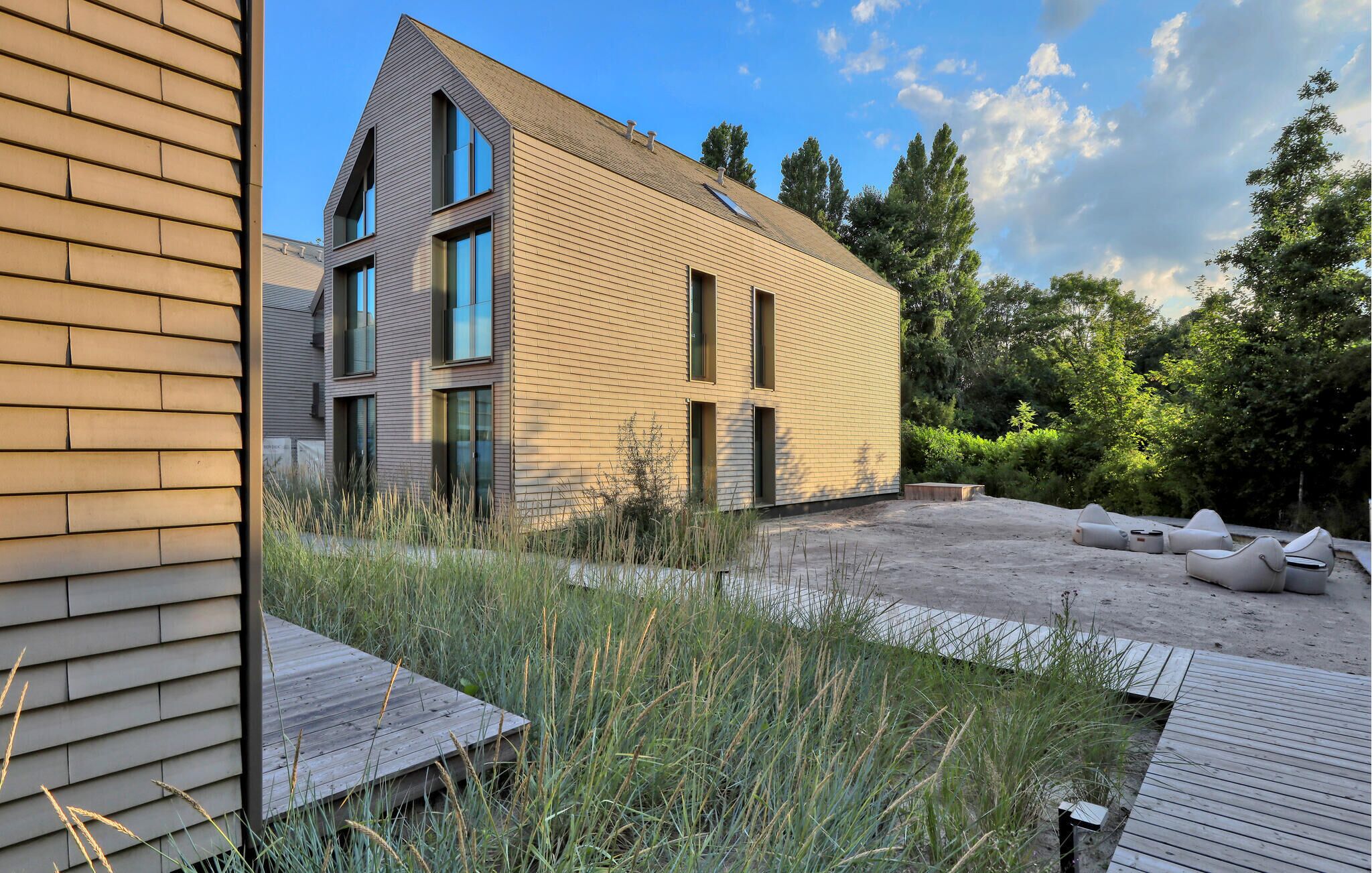 A modern two-story building with a sloped roof and large windows, surrounded by a landscaped garden with a wooden deck.