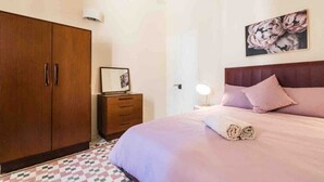 A bedroom with a large wooden wardrobe, a bedside table with a lamp, a bed with a headboard, and a framed picture on the wall. - Cottonera cara guesthouse (Bormla)