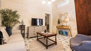 A living room with a sofa, a coffee table, a television, a wooden cabinet, and a large plant. - Cottonera cara guesthouse (Bormla)