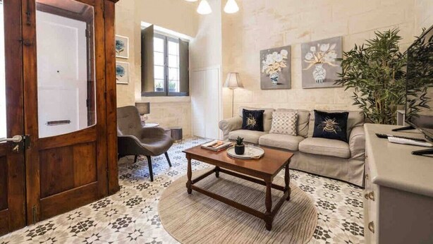 A living room with a wooden door, a sofa set, a coffee table, and a patterned rug. - Cottonera cara guesthouse (Bormla)