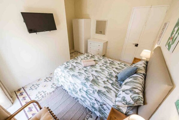 Ein Schlafzimmer mit einem Bett, einem an der Wand montierten Fernseher, einem Nachttisch mit Lampe und einer Tür.