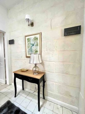 Un couloir avec un sol carrelé, une petite table sur laquelle se trouve une lampe, un tableau encadré et une pancarte indiquant « Bermudes ».