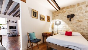 A bedroom with a stone wall, a bed with white bedding and a red pillow, a wooden chair, and a small table with a lamp.