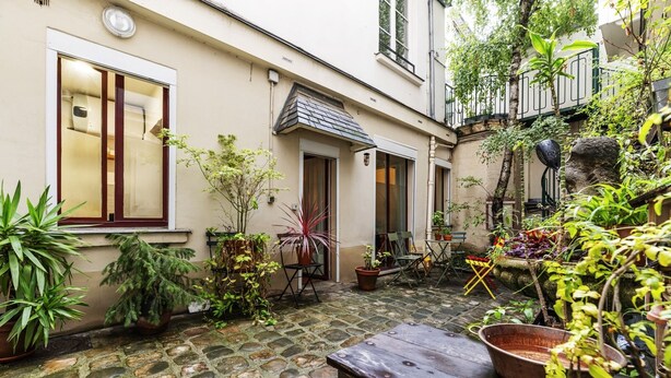 A courtyard with stone flooring, potted plants, a wooden table, and a stone statue.