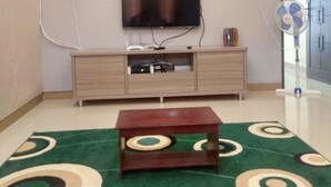 Un salon avec un téléviseur à écran plat fixé au mur, une table basse en bois et un tapis à motifs verts.