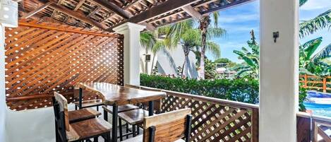 Un patio al aire libre con mesas y sillas de madera, un muro de celosía de madera y vistas a una piscina y palmeras.