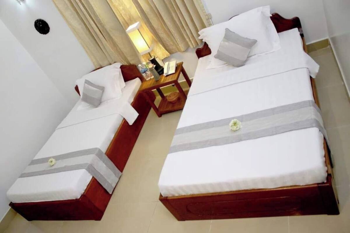 Une chambre d’hôtel avec deux lits simples, une petite table en bois, une lampe et une fenêtre avec des rideaux.