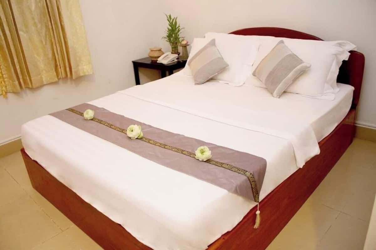 Un lit bien fait, avec du linge de lit blanc et des coussins décoratifs, une table de chevet avec un téléphone et une petite plante, et une fenêtre avec des rideaux.