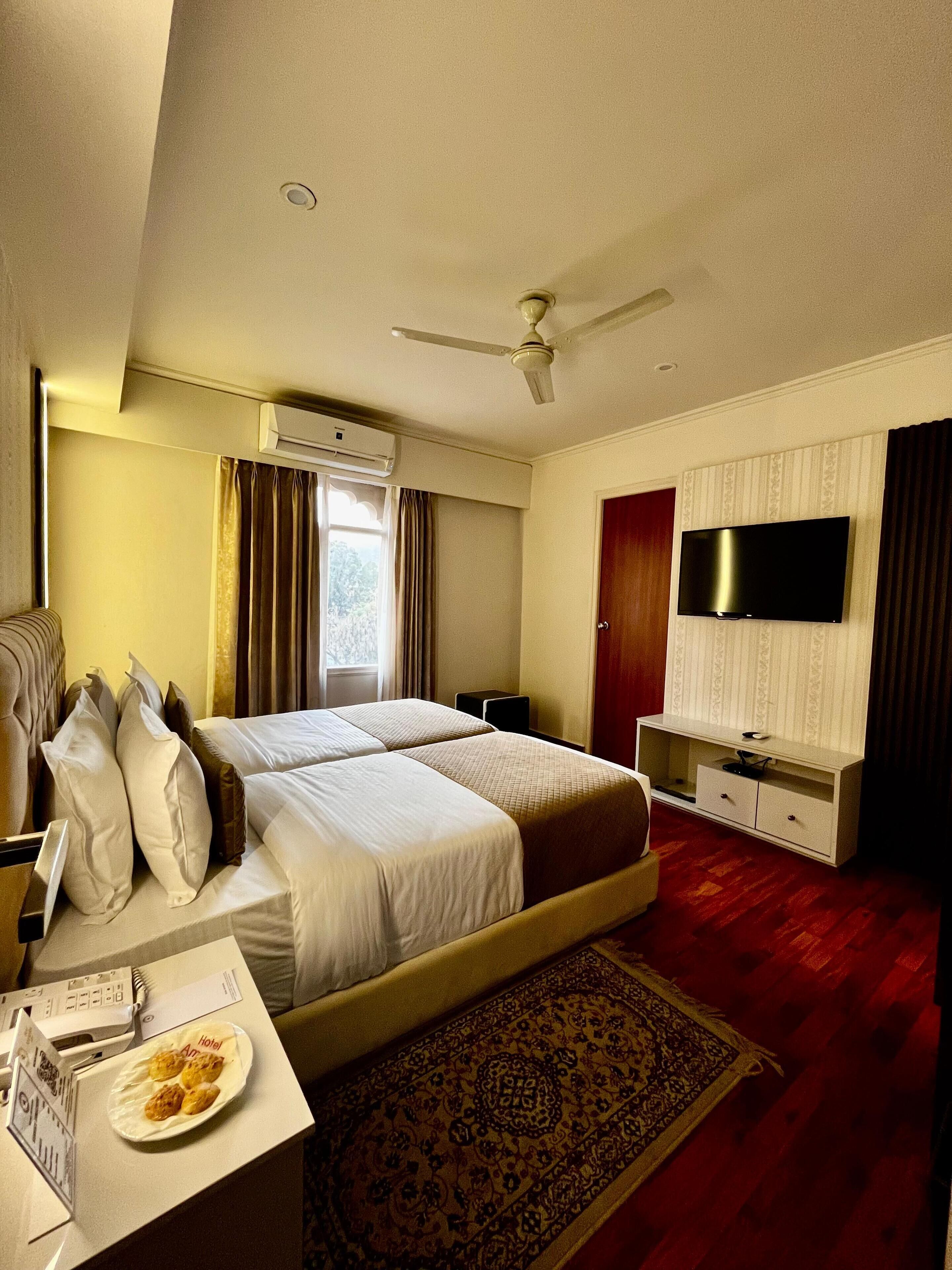A hotel room with two beds, a flat-screen TV, an air conditioning unit, a desk with a plate of food, and a patterned rug.