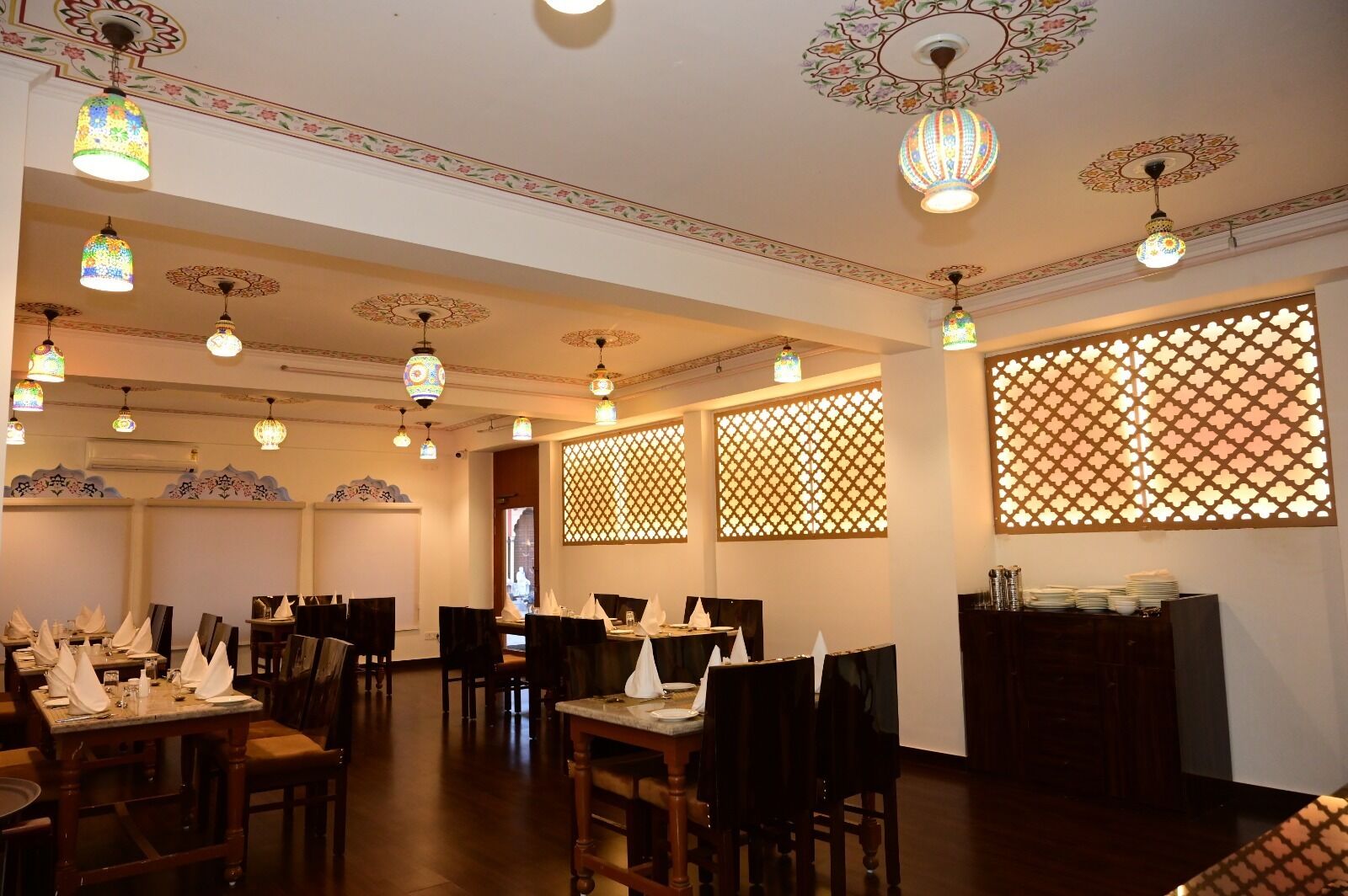 Un comedor con mesas dispuestas para el servicio, lámparas de techo ornamentadas y paneles decorativos en la pared.