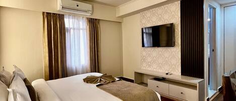 A hotel room with a large bed, an air conditioning unit, a ceiling fan, a flat-screen TV mounted on the wall, a wooden floor, and a desk with a chair.