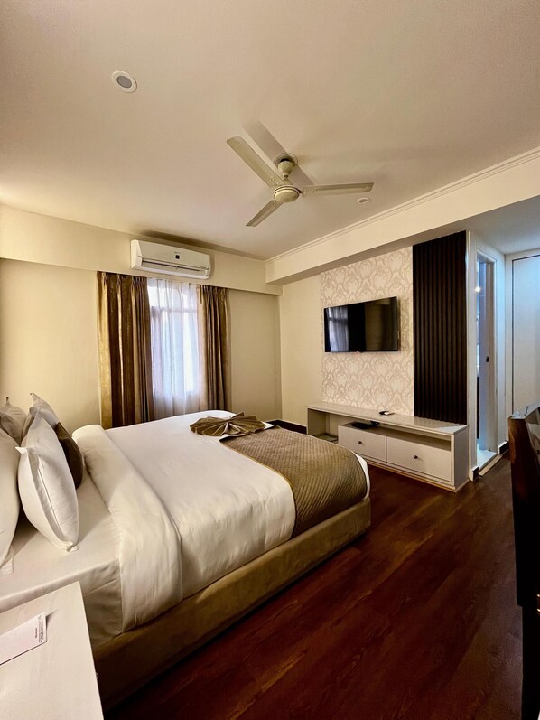 Habitación de hotel con una cama grande, un equipo de aire acondicionado, un ventilador de techo, un televisor de pantalla plana montado en la pared, piso de madera y un escritorio con una silla.