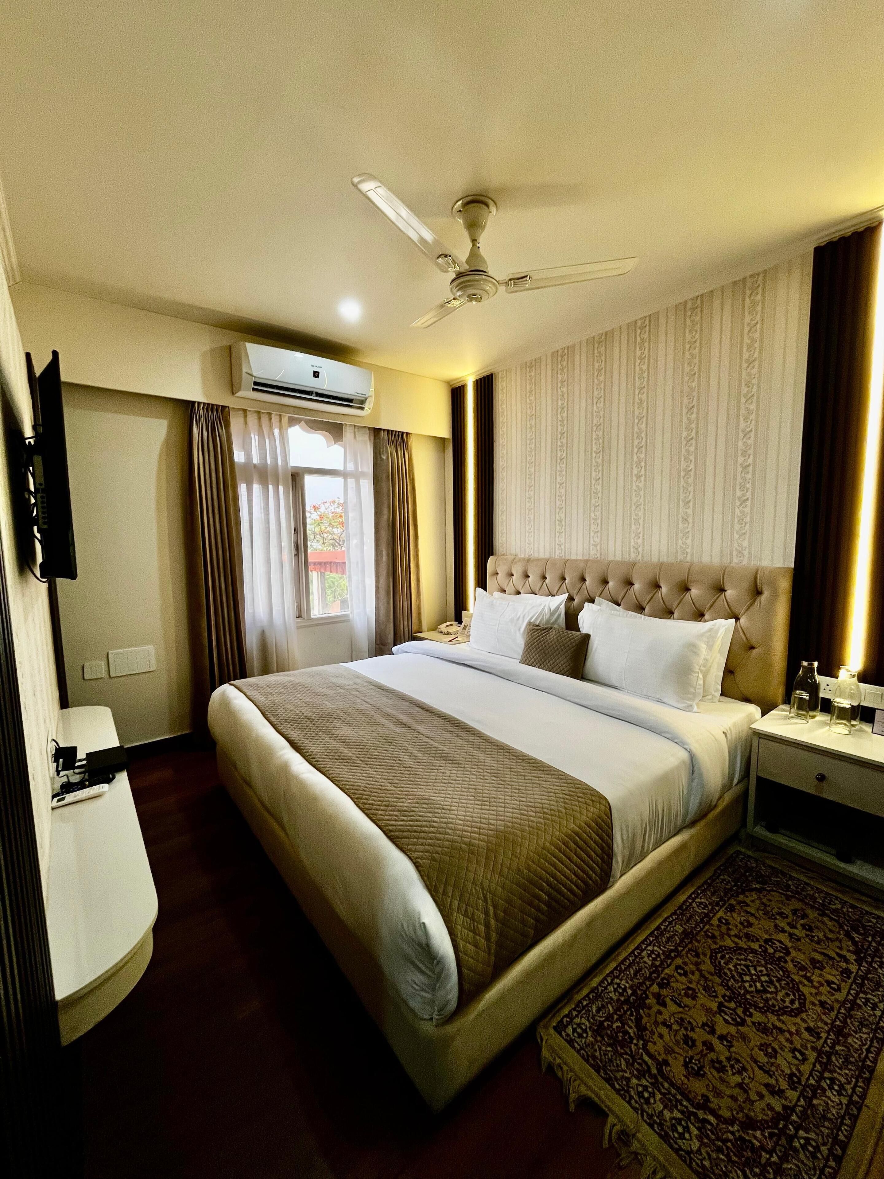 A hotel room with a large bed, a ceiling fan, an air conditioning unit, a bedside table with a lamp, and a patterned rug.