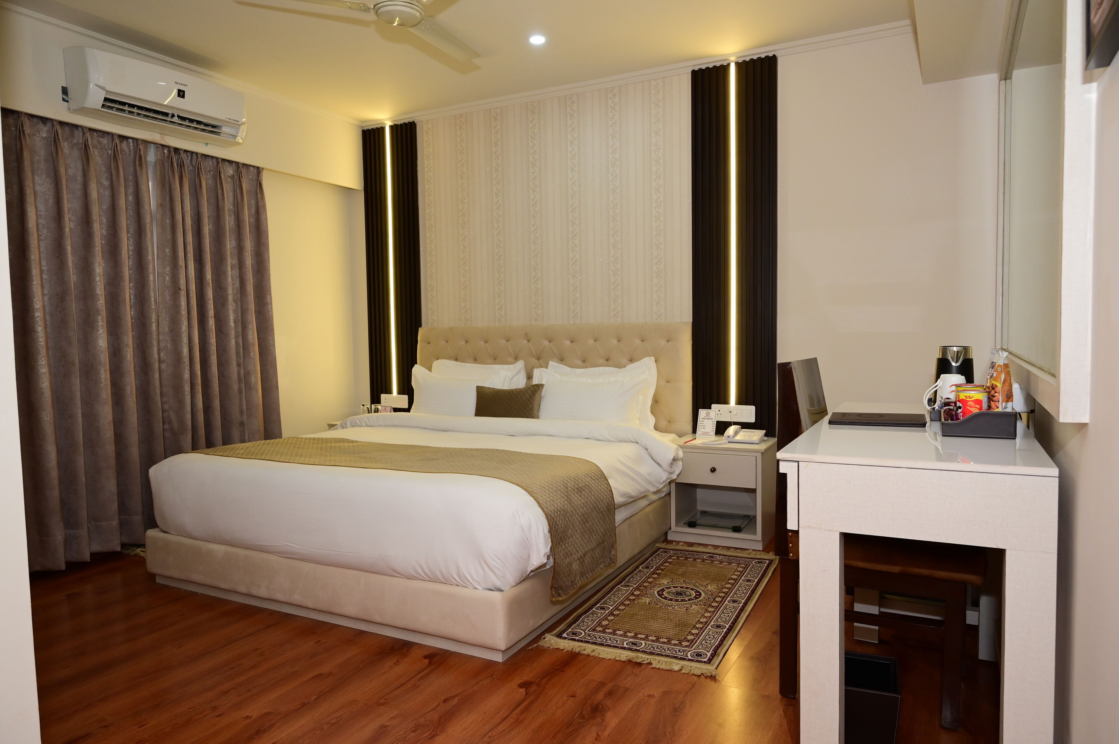 Una habitación de hotel con una cama grande, una mesita de noche con lámpara, un equipo de aire acondicionado y un pequeño escritorio con espejo y artículos de aseo.