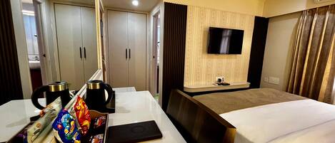 Habitación de hotel con cama, escritorio con hervidor, cafetera, ordenador portátil y televisor montado en la pared.