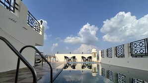 Una piscina en la azotea con vistas despejadas del cielo y nubes, rodeada por un edificio blanco con barandillas decoradas.