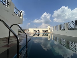 Piscina no terraço com vista ampla do céu e nuvens, cercada por um edifício branco com grades estampadas.