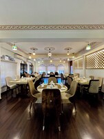 Um restaurante com várias mesas e cadeiras, medalhões decorativos no teto e vista para a rua através de grandes janelas.