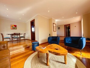 A living room with a wooden coffee table, blue armchairs, a brown sofa, a dining table with chairs, and a blue trash bin. - Mountains and Treetops Retreat - Nature Getaway (West Moonah)
