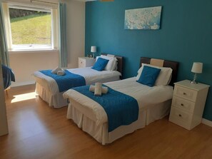 Two single beds with blue and white bedspreads, a window with a view of a grassy area, and a bedside table with a lamp.
