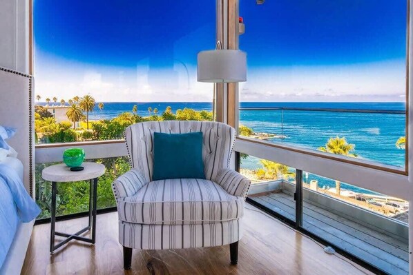 Une pièce dotée d’une grande fenêtre donnant sur une plage, d’un fauteuil à rayures, d’une petite table surmontée d’un vase vert et d’un luminaire suspendu.