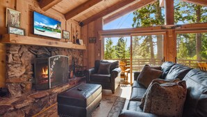 A cozy cabin interior with a stone fireplace, leather furniture, and a large window offering a view of the forest. - Sweet Tahoe Cabin, Lake Views/Hot Tub/Secluded (Carnelian Bay)