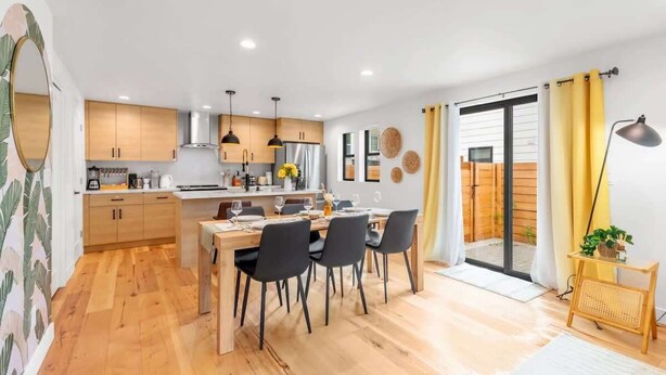 A modern kitchen with wooden cabinets, a dining area with black chairs, and a sliding glass door leading to a balcony.