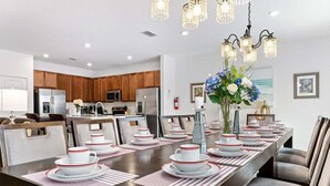 A dining area with a long table set for a meal, kitchen in the background, and a chandelier above. - Spacious 10BR resort home with private pool, game room, and room for 25. (Kissimmee)