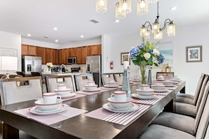 A dining area with a long table set for a meal, kitchen in the background, and a chandelier above. - Spacious 10BR resort home with private pool, game room, and room for 25. (Kissimmee)