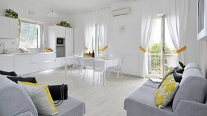 Una sala de estar moderna y bien iluminada con un sofá gris, un comedor con mesa y sillas, y una cocina con gabinetes y electrodomésticos blancos.