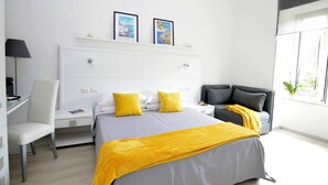 Una habitación de hotel moderna con una cama, mesitas de noche, un escritorio, una silla y un sofá. Hay dos cuadros enmarcados en la pared sobre la cama.