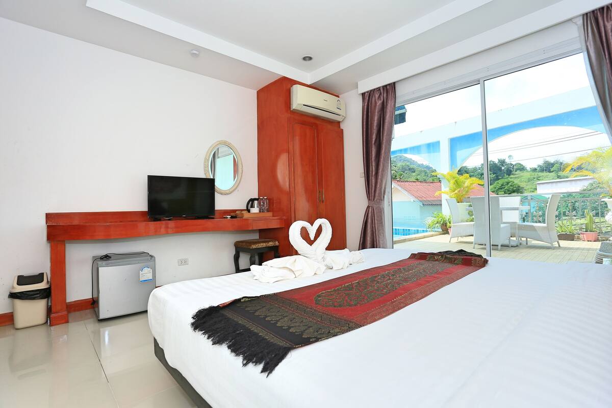 A hotel room with a bed, a TV, a small fridge, a desk with a mirror, and a view of the outdoors.