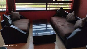 Una habitación con dos sofás y una mesa de centro, ventanas amplias que dan a un edificio y zonas verdes.