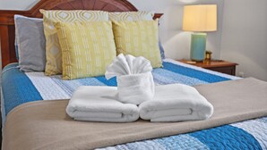 Una cama bien tendida con un edredón azul y blanco, dos almohadas estampadas y una toalla doblada sobre una manta color beige.