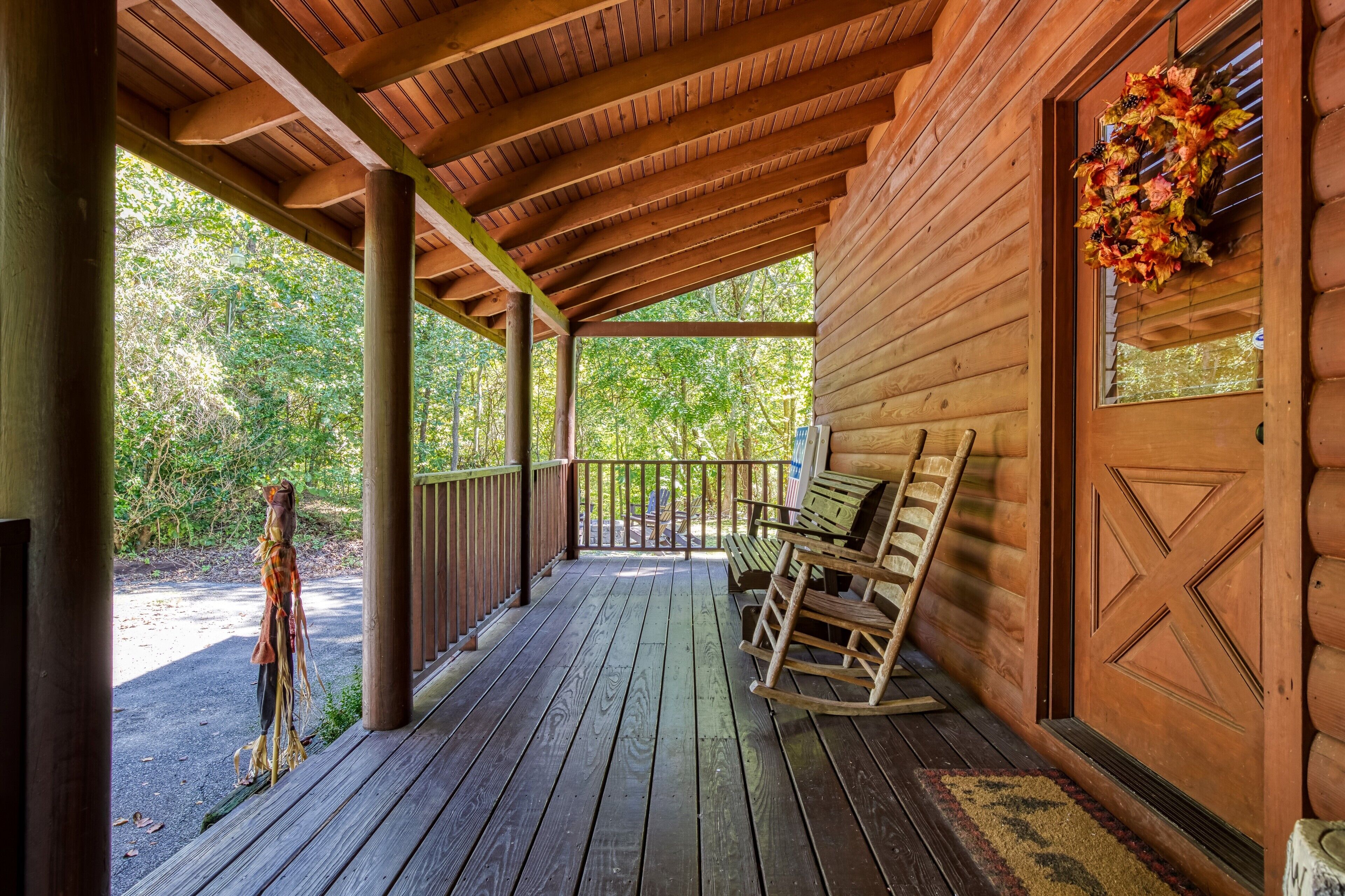 A wooden porch with a rocking chair and a bench, a wooden door with a decorative wreath, and a view of a forested area.