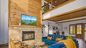 A modern living room with a stone fireplace, a television mounted on a wooden wall, and a blue leather sofa. - A Piece Of Perfect: Pool, Mtn Views, Home Theater, Game Room, Hot Tub, Wifi (Sevierville)