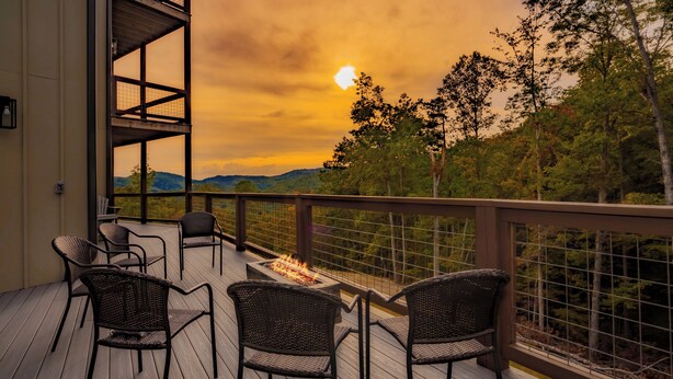 A deck with wicker chairs and a fire feature, overlooking a forested area with a sunset view. - A Piece Of Perfect: Pool, Mtn Views, Home Theater, Game Room, Hot Tub, Wifi (Sevierville)