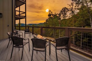 Una terraza con sillas de mimbre y una chimenea, con vistas a una zona boscosa y una puesta de sol.