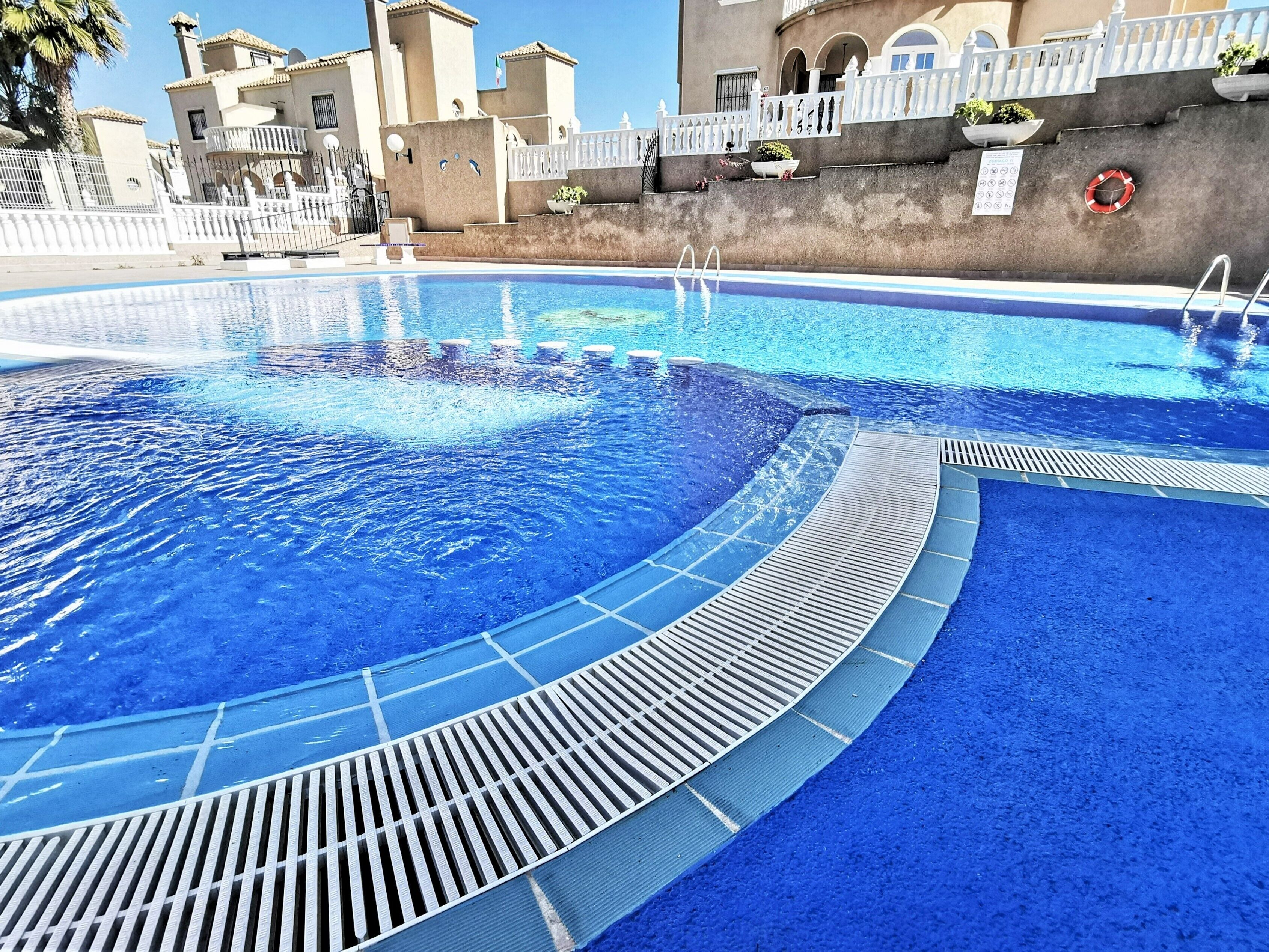A swimming pool with a clear blue water feature, surrounded by a white fence and a building with balconies.