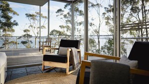 A modern room with large windows, a wooden deck, and a view of the sea and trees.