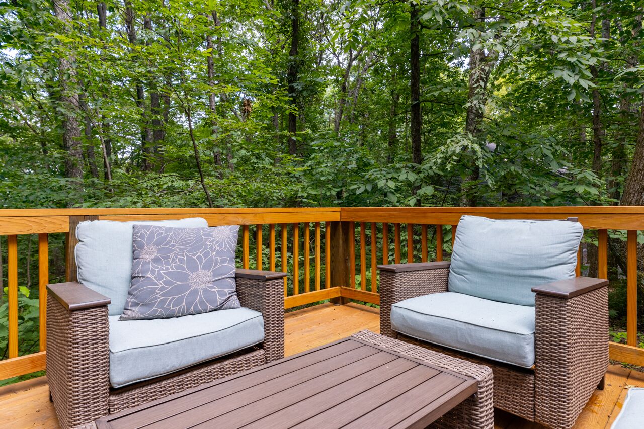 A wooden deck with wicker furniture and a table, surrounded by dense green foliage.