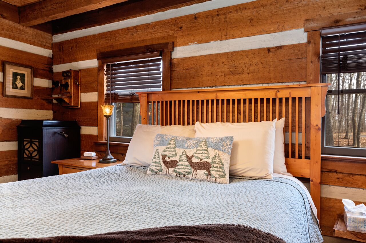 A cozy log cabin bedroom with a wooden headboard, a bed with a white and brown blanket, a decorative pillow with a forest scene, a nightstand with a lamp, and a window with blinds.