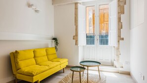 Ein helles Wohnzimmer mit einem gelben Sofa, einem Glastisch und einem gemusterten Teppich.