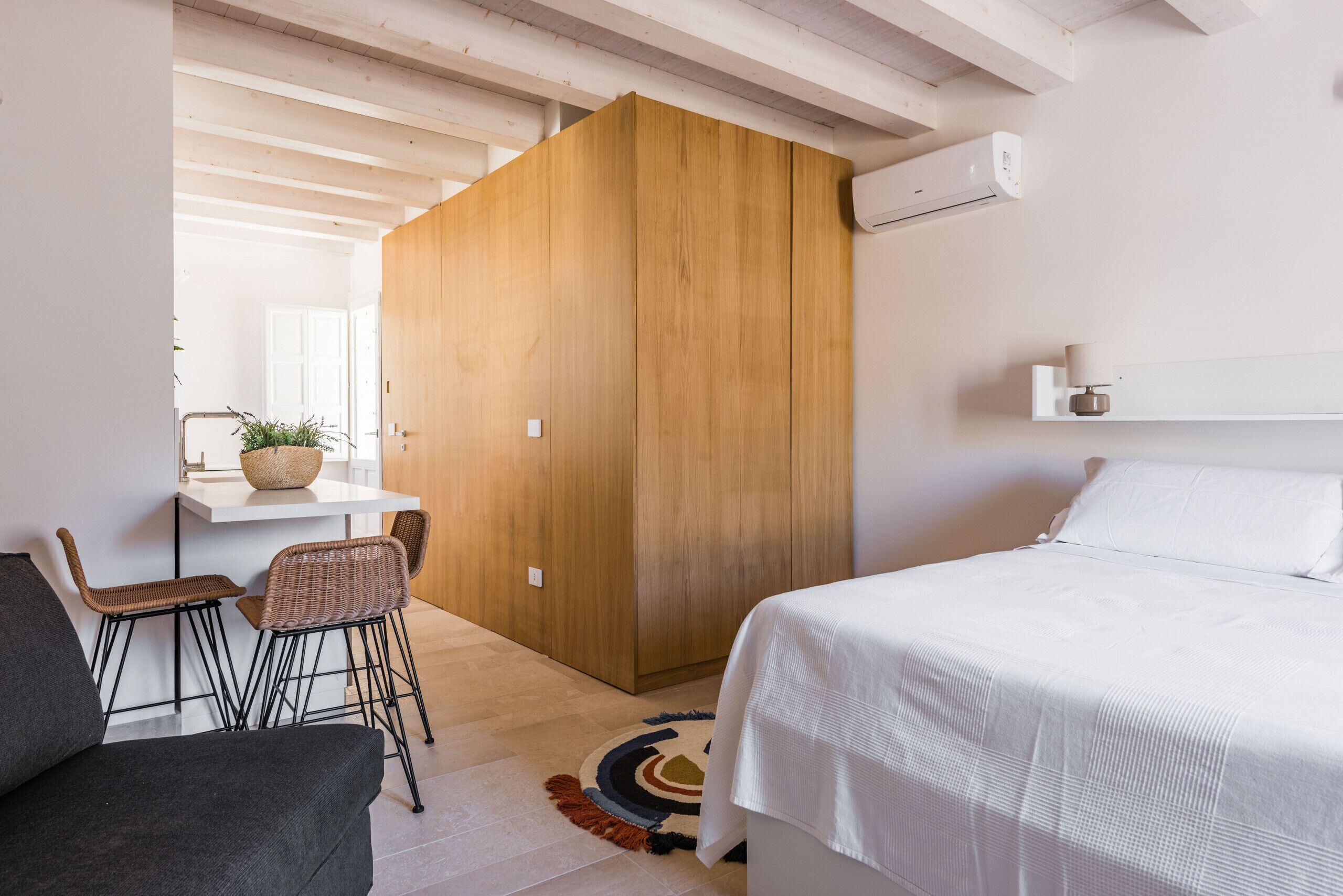 A modern bedroom with a large bed, a wooden wardrobe, a small table with chairs, and a potted plant.