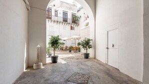 Un corredor arqueado con plantas en macetas, una fuente y vistas a un edificio con balcones y mobiliario exterior.
