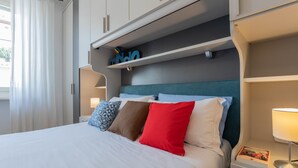 Letto rifatto con lenzuola bianche e numerosi cuscini di diversi colori. La testata presenta un dettaglio color petrolio e, sopra il letto, si trova una mensola con oggetti decorativi.