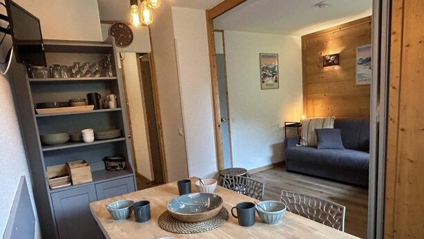 Een eethoek met een houten tafel voor vier personen, omringd door stoelen. Links is een keukenruimte met planken en een klok te zien.