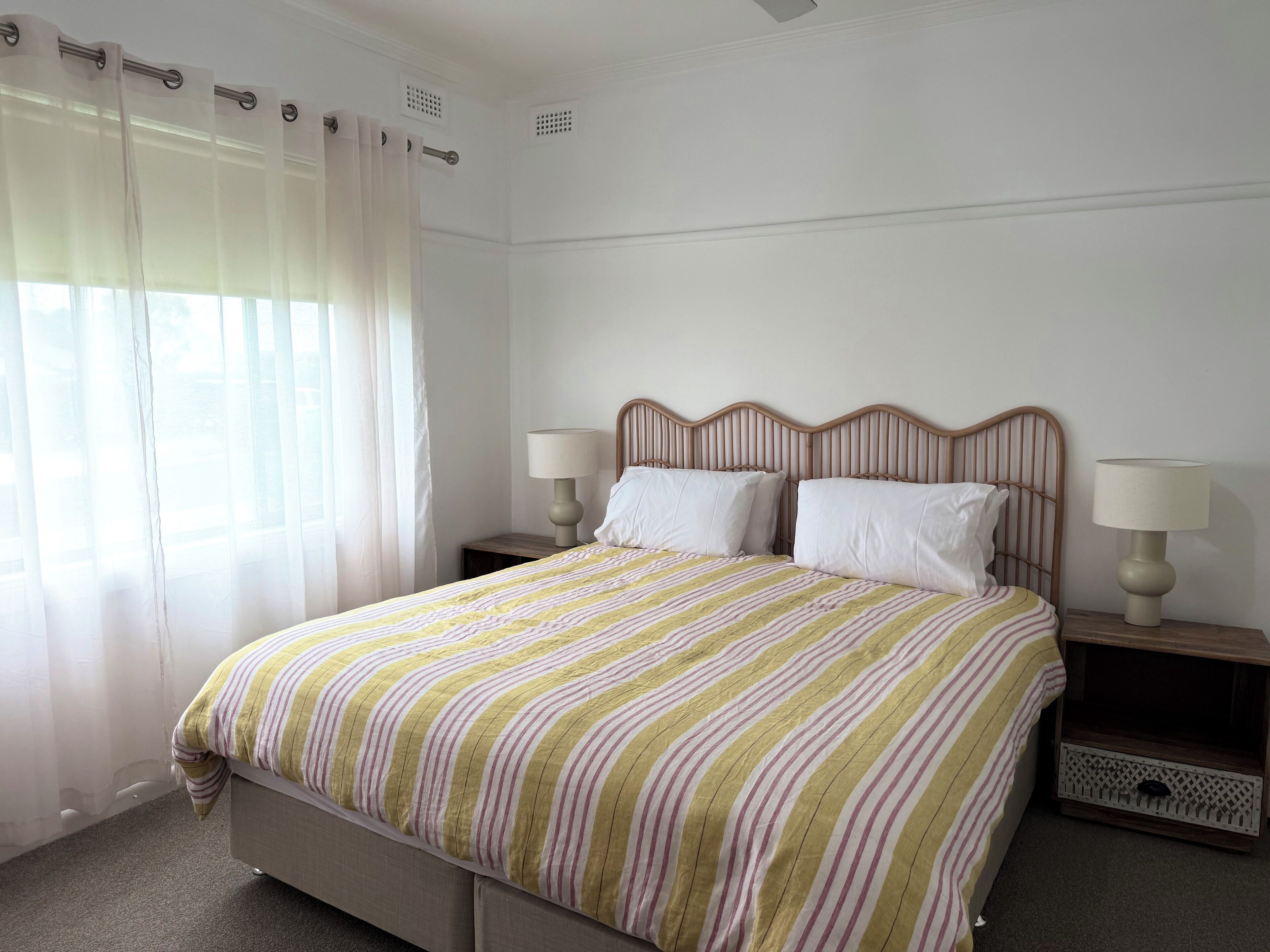 A neatly made bed with a striped bedspread, two bedside tables with lamps, and a window with sheer curtains.